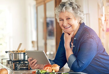 Mujer mayor sonriente con tableta en la cocina, rodeada de vegetales frescos y una olla sobre la estufa, mostrando hábitos activos y alimentación en el adulto mayor