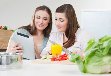 Dos amigas en una cocina junto a vegetales picados en una tabla, preparando una comida saludable para su práctica de ayuno intermitente