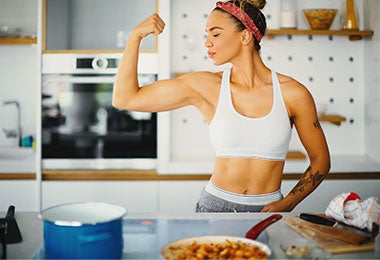 Mujer atlética con top blanco y pantalones grises, mostrando su bíceps en una cocina, lista para preparar alimento para ganar masa muscular