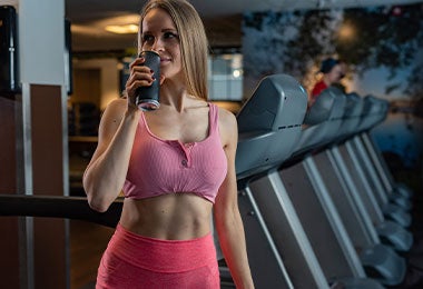 Mujer sonriente con ropa deportiva rosa, de pie en un gimnasio junto a las cintas de correr, tomando una bebida como alimento para ganar masa muscular