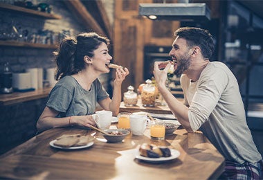 Una pareja sonriente sentada en una barra de cocina, comiendo y riendo mientras disfrutan de su desayuno durante el ayuno intermitente