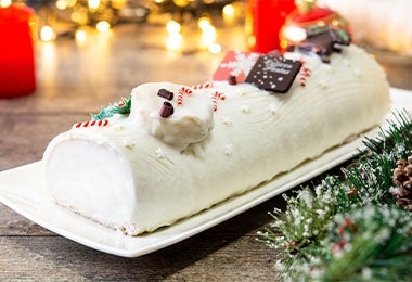 Pastel de Navidad tipo tronco blanco, decorado con bastones de caramelo, disco de chocolate y ramas de pino, ideal para celebraciones festivas sobre una mesa con luces de fondo