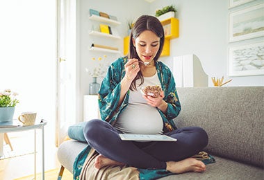 Mujer embarazada sentada sobre la cama con las piernas cruzadas, sonriendo mientras come una ensalada, reflejo de una alimentación en el embarazo