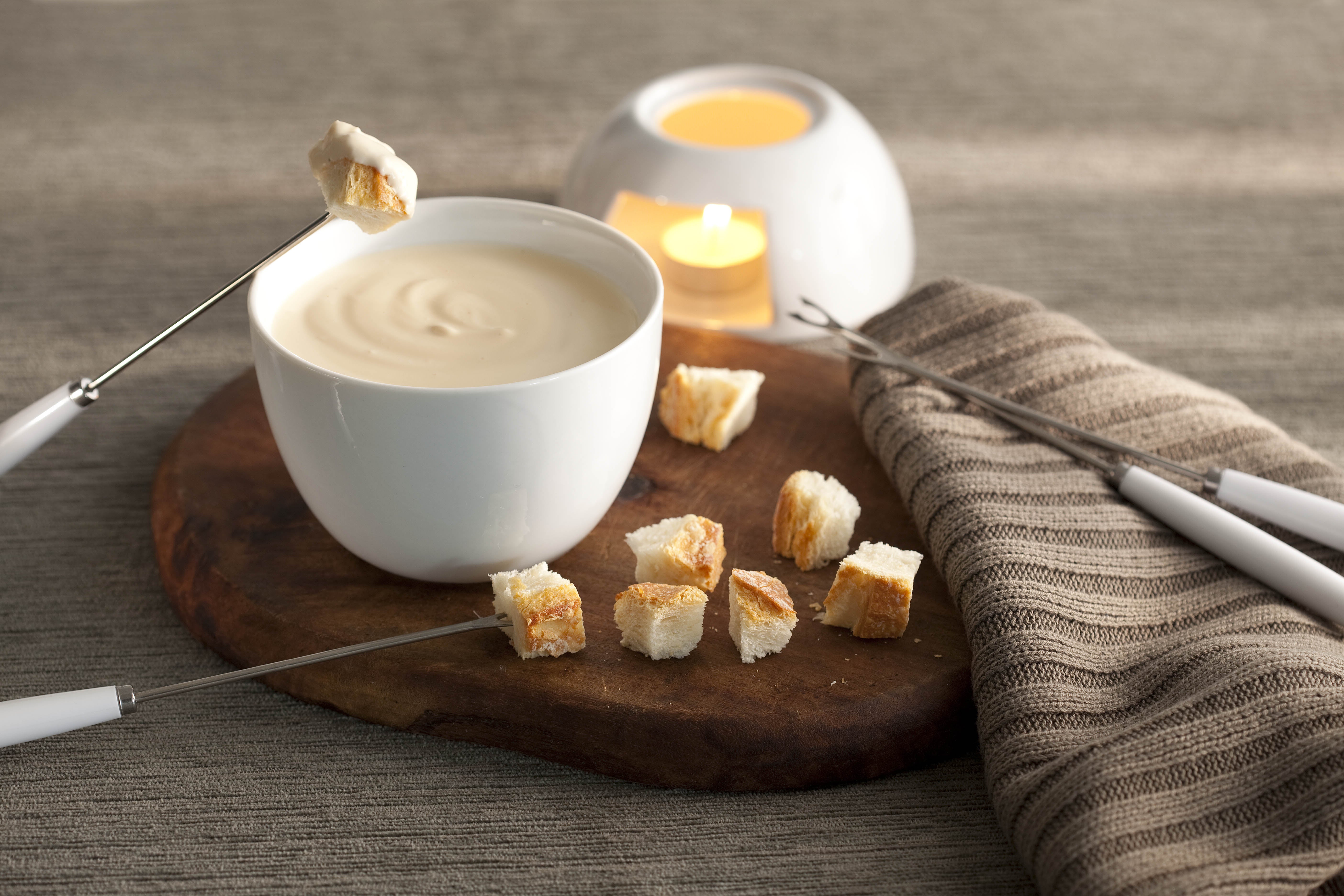 fotografía en tonos grises, marrones y banco visto de frente. Contiene una tabla de madera redonda con un recipiente blanco redondo que contiene la fondue, 4 tenedores de fondue y trozos de pan alrededor.