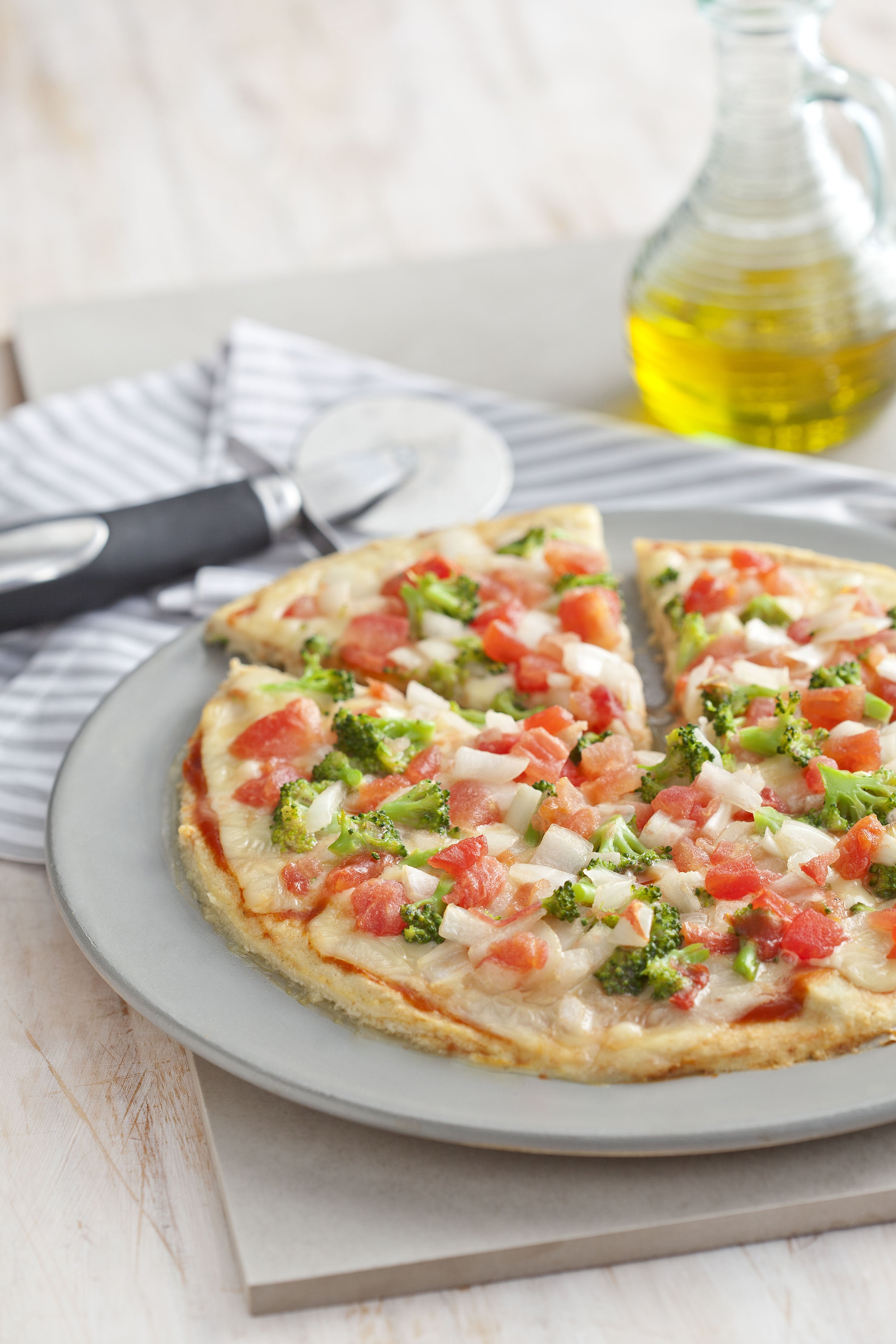 La fotografía en tonos blancos de una encimera de vista frontal contiene un plato blanco redondo con una pizza con trozos de tomate y brócoli en el fondo y una jarra de aceite.