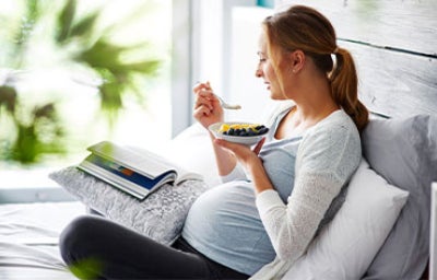 Mujer embarazada recostada en la cama, disfrutando un tazón de frutas mientras lee, ejemplo de alimentación en el embarazo