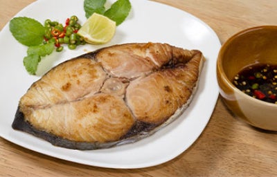 Un filete de pescado dorado y jugoso servido en un plato blanco con rodajas de limón y hojas frescas