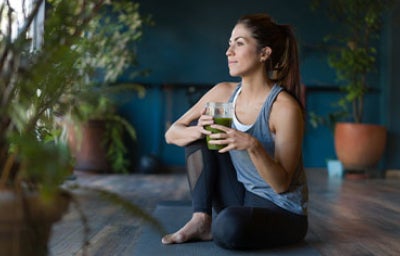Mujer sentada en una esterilla de yoga, con un batido verde en la mano, ideal como alimento para ganar masa muscular
