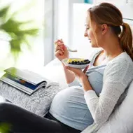 Mujer embarazada recostada en la cama, disfrutando un tazón de frutas mientras lee, ejemplo de alimentación en el embarazo
