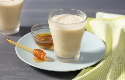 Fotografía en tonos amarillos con fondo gris con plato blanco redondo celeste, tarro de miel y pinzas, un vaso de vidrio con la bebida y un paño a cuadros verde y blanco.