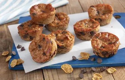 Fotografía en tonos de azul en un banco de madera con un tablero azul, con una hoja de papel encerado y varios muffins de plátano con chispas de chocolate encima. Cereales para el desayuno Nesfit y algunas chispas de chocolate esparcidas por el mostrador.