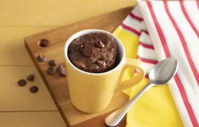 Fotografía en tonos amarillos sobre un banco de madera amarillo, una tela a rayas rojas y amarillas y una tabla de madera en el centro de la foto con una taza amarilla con el pastel de chocolate descansando sobre ella. Fotografía en tonos amarillos sobre un banco de madera amarillo, una tela a rayas rojas y amarillas y una tabla de madera en el centro de la foto con una taza amarilla con el pastel de chocolate descansando sobre ella.