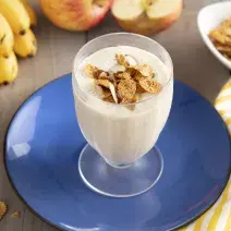 Imagen de un vaso con bebida cremosa en tono claro, decorado con cereal. Está en un plato azul, y alrededor hay cereales en la mesa, un racimo de plátanos, dos manzanas y una tela de rayas amarillas y blancas.