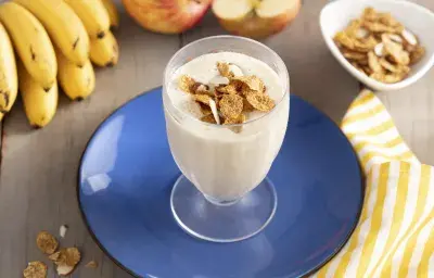 Imagen de un vaso con bebida cremosa en tono claro, decorado con cereal. Está en un plato azul, y alrededor hay cereales en la mesa, un racimo de plátanos, dos manzanas y una tela de rayas amarillas y blancas. Imagen de un vaso con bebida cremosa en tono claro, decorado con cereal. Está en un plato azul, y alrededor hay cereales en la mesa, un racimo de plátanos, dos manzanas y una tela de rayas amarillas y blancas.