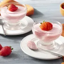 Imagen de primer plano de la receta de Strawberry Mousse Zero Lactose, en rosa claro, servida en frascos de vidrio, decorada con rodajas de fresas. La receta está en platos blancos sobre un mostrador con una tela naranja y más fresas enteras decorando