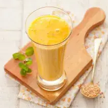 Fotografía en tonos amarillos y blancos tomada desde un banco visto de frente, contiene una tabla de madera y un vaso transparente con una bebida de mango encima.