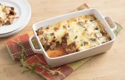 Fotografía en tonos blancos y amarillos de encimeras de madera clara, con tela a cuadros rojos y verdes, fuente rectangular blanca con asas y trozos de berenjena y queso en su interior. Al fondo, tres platos blancos apilados con un trozo de la receta.