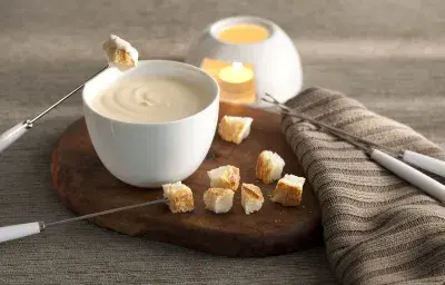 fotografía en tonos grises, marrones y banco visto de frente. Contiene una tabla de madera redonda con un recipiente blanco redondo que contiene la fondue, 4 tenedores de fondue y trozos de pan alrededor.