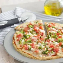 La fotografía en tonos blancos de una encimera de vista frontal contiene un plato blanco redondo con una pizza con trozos de tomate y brócoli en el fondo y una jarra de aceite.