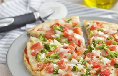 La fotografía en tonos blancos de una encimera de vista frontal contiene un plato blanco redondo con una pizza con trozos de tomate y brócoli en el fondo y una jarra de aceite.