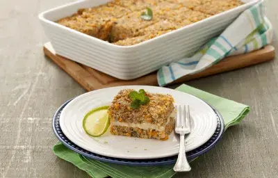 Fotografía en tonos de verde sobre un banco de madera gris, una tabla de cortar de madera, un paño de cocina verde, uno rayado en verde. En el centro, un plato redondo blanco con un trozo de kebab relleno. Al fondo, un refractario con toda la kibbeh. Fotografía en tonos de verde sobre un banco de madera gris, una tabla de cortar de madera, un paño de cocina verde, uno rayado en verde. En el centro, un plato redondo blanco con un trozo de kebab relleno. Al fondo, un refractario con toda la kibbeh.