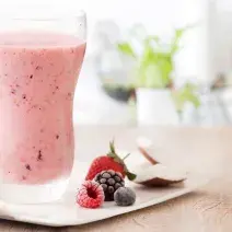 fotografía y tonos de rosa y blanco de un banco beige visto de frente, contiene un vaso transparente con la bebida de frutos rojos y al lado contiene frutos del bosque.