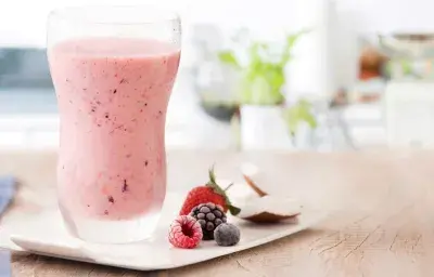 fotografía y tonos de rosa y blanco de un banco beige visto de frente, contiene un vaso transparente con la bebida de frutos rojos y al lado contiene frutos del bosque.