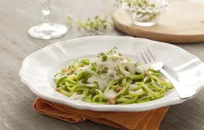 Fotografía en tonos verdes y blancos, en el centro un plato blanco profundo con espaguetis de calabacín verde en el interior con salsa blanca. Debajo del plato hay una servilleta de tela naranja sobre una mesa de madera.