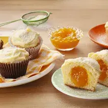 Fotografia em tons de laranja em uma bancada de madeira com um prato branco com paninho listrado em amarelo e três cupcakes de leite Ninho com manga em cima dele. Ao lado, um prato branco com um cupcake cortado ao meio mostrando o recheio de manga.