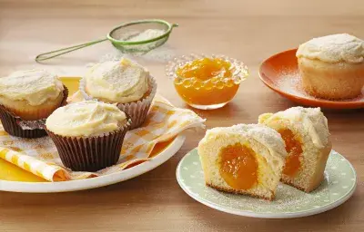 Fotografia em tons de laranja em uma bancada de madeira com um prato branco com paninho listrado em amarelo e três cupcakes de leite Ninho com manga em cima dele. Ao lado, um prato branco com um cupcake cortado ao meio mostrando o recheio de manga.