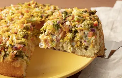 Fotografía amarilla y blanca de un plato amarillo con pastel de verduras y una espátula quitando una rebanada. Junto a él hay una tela blanca.