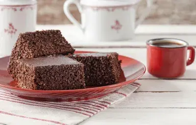 Fotografía en tonos rojos sobre una encimera de madera clara con un paño rayado en blanco y rojo y un plato rojo con tres piezas de bizcocho de cacao con coco. A su lado, una taza de café roja y al fondo, dos teteras blancas.