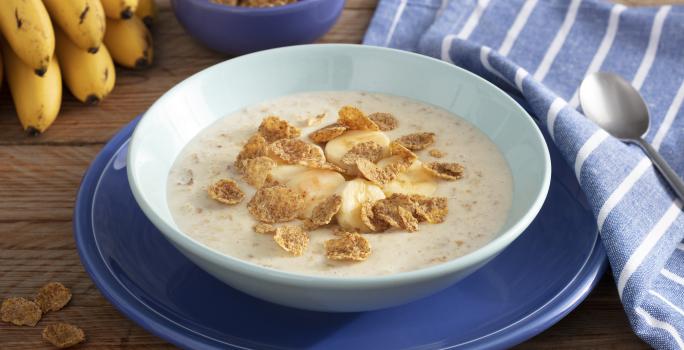 Fotografía en tonos de azul sobre un banco de madera con un paño azul con rayas blancas, un gran plato azul en el centro, con un plato hondo celeste y la papilla con cereales. Al fondo, un racimo de plátanos y una pequeña olla de cereal.