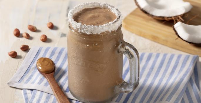 Paño rayado celeste y blanco con cuchara de madera marrón con mantequilla de maní. Una taza transparente con el batido y el borde de coco rallado.