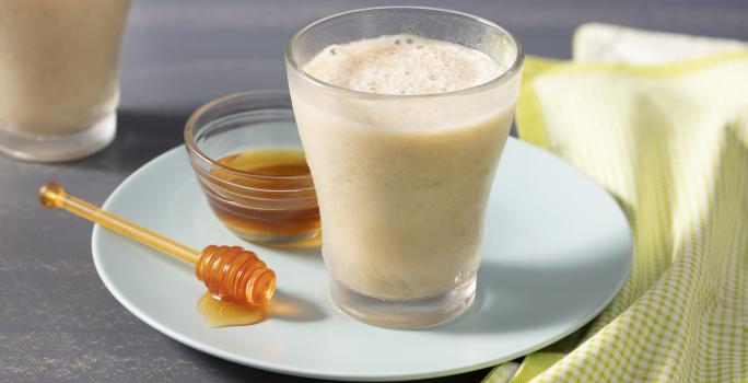 Fotografía en tonos amarillos con fondo gris con plato blanco redondo celeste, tarro de miel y pinzas, un vaso de vidrio con la bebida y un paño a cuadros verde y blanco.