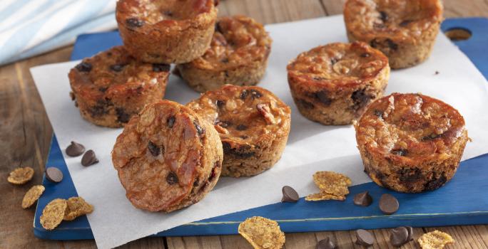 Fotografía en tonos de azul en un banco de madera con un tablero azul, con una hoja de papel encerado y varios muffins de plátano con chispas de chocolate encima. Cereales para el desayuno Nesfit y algunas chispas de chocolate esparcidas por el mostrador.