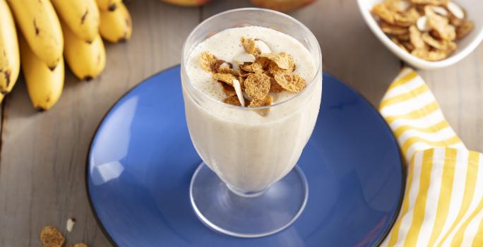 Imagen de un vaso con bebida cremosa en tono claro, decorado con cereal. Está en un plato azul, y alrededor hay cereales en la mesa, un racimo de plátanos, dos manzanas y una tela de rayas amarillas y blancas.