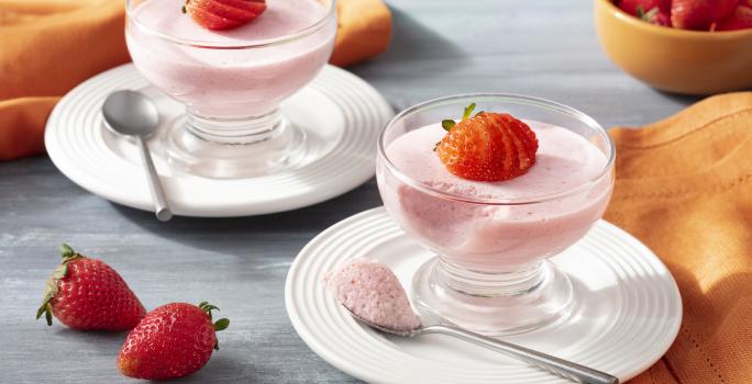 Imagen de primer plano de la receta de Strawberry Mousse Zero Lactose, en rosa claro, servida en frascos de vidrio, decorada con rodajas de fresas. La receta está en platos blancos sobre un mostrador con una tela naranja y más fresas enteras decorando