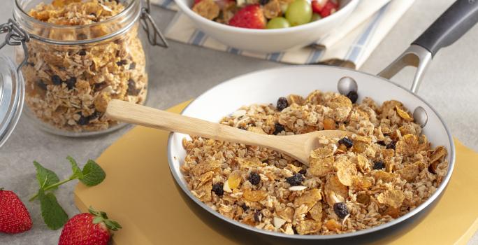 Fotografía en tonos amarillos en un banco de madera con una tabla de cortar de madera, una gran sartén adentro con la granola con pasas, cereal de desayuno adentro. Al lado, una olla pequeña con la granola y al lado otra olla blanca con fruta.