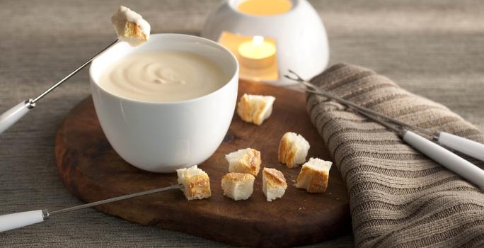 fotografía en tonos grises, marrones y banco visto de frente. Contiene una tabla de madera redonda con un recipiente blanco redondo que contiene la fondue, 4 tenedores de fondue y trozos de pan alrededor.