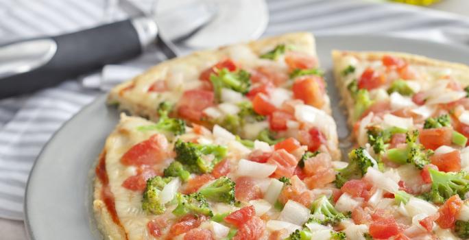 La fotografía en tonos blancos de una encimera de vista frontal contiene un plato blanco redondo con una pizza con trozos de tomate y brócoli en el fondo y una jarra de aceite.