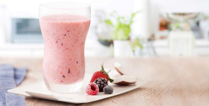 fotografía y tonos de rosa y blanco de un banco beige visto de frente, contiene un vaso transparente con la bebida de frutos rojos y al lado contiene frutos del bosque.