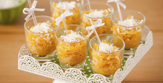 Fotografía en tonos naranjas sobre una mesa de madera con un puesto de caramelos blancos en el centro con caramelos de calabaza en vasos transparentes con palillos chinos y un lazo blanco.