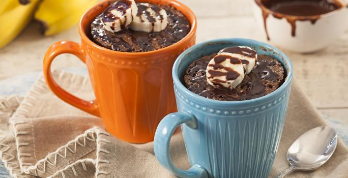 Fotografía en tonos beige, azul y naranja de una encimera blanca con un paño beige, sobre ella una taza azul y una taza naranja con pastel de taza. En el fondo un racimo de plátanos y un recipiente redondo con almíbar.