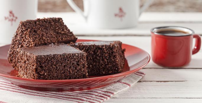 Fotografía en tonos rojos sobre una encimera de madera clara con un paño rayado en blanco y rojo y un plato rojo con tres piezas de bizcocho de cacao con coco. A su lado, una taza de café roja y al fondo, dos teteras blancas.