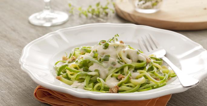 Fotografía en tonos verdes y blancos, en el centro un plato blanco profundo con espaguetis de calabacín verde en el interior con salsa blanca. Debajo del plato hay una servilleta de tela naranja sobre una mesa de madera.