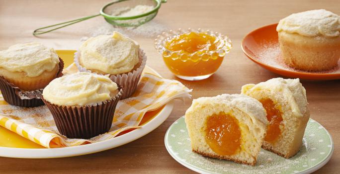 Fotografia em tons de laranja em uma bancada de madeira com um prato branco com paninho listrado em amarelo e três cupcakes de leite Ninho com manga em cima dele. Ao lado, um prato branco com um cupcake cortado ao meio mostrando o recheio de manga.