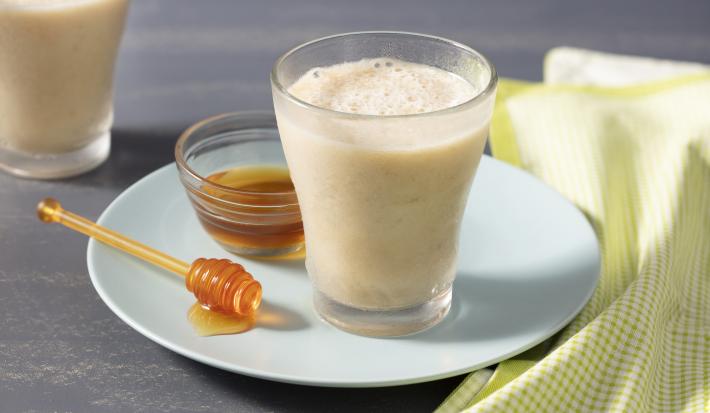 Fotografía en tonos amarillos con fondo gris con plato blanco redondo celeste, tarro de miel y pinzas, un vaso de vidrio con la bebida y un paño a cuadros verde y blanco.