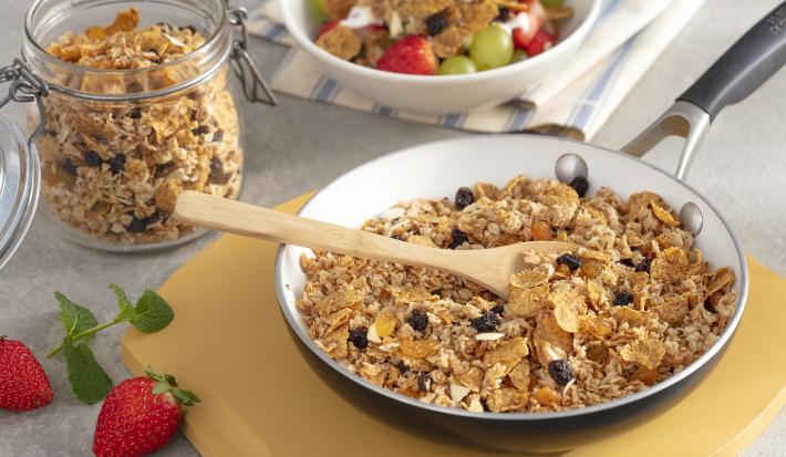 Fotografía en tonos amarillos en un banco de madera con una tabla de cortar de madera, una gran sartén adentro con la granola con pasas, cereal de desayuno adentro. Al lado, una olla pequeña con la granola y al lado otra olla blanca con fruta.