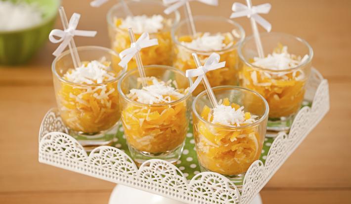 Fotografía en tonos naranjas sobre una mesa de madera con un puesto de caramelos blancos en el centro con caramelos de calabaza en vasos transparentes con palillos chinos y un lazo blanco.